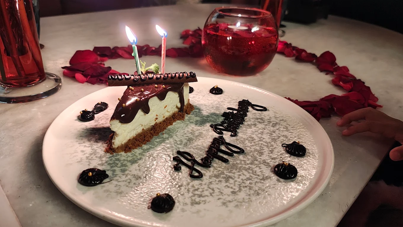 A birthday cheesecake slice with two lit candles, rose petals around the plate.