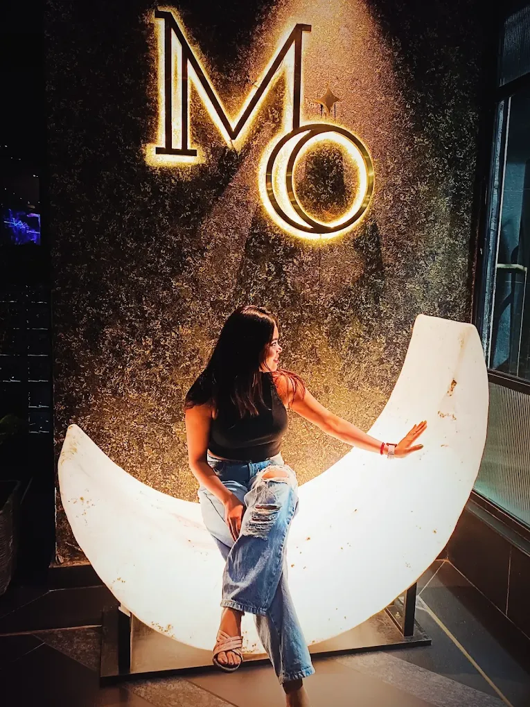 The glowing crescent-moon seat under the MO sign, a guest sitting in its curve.
