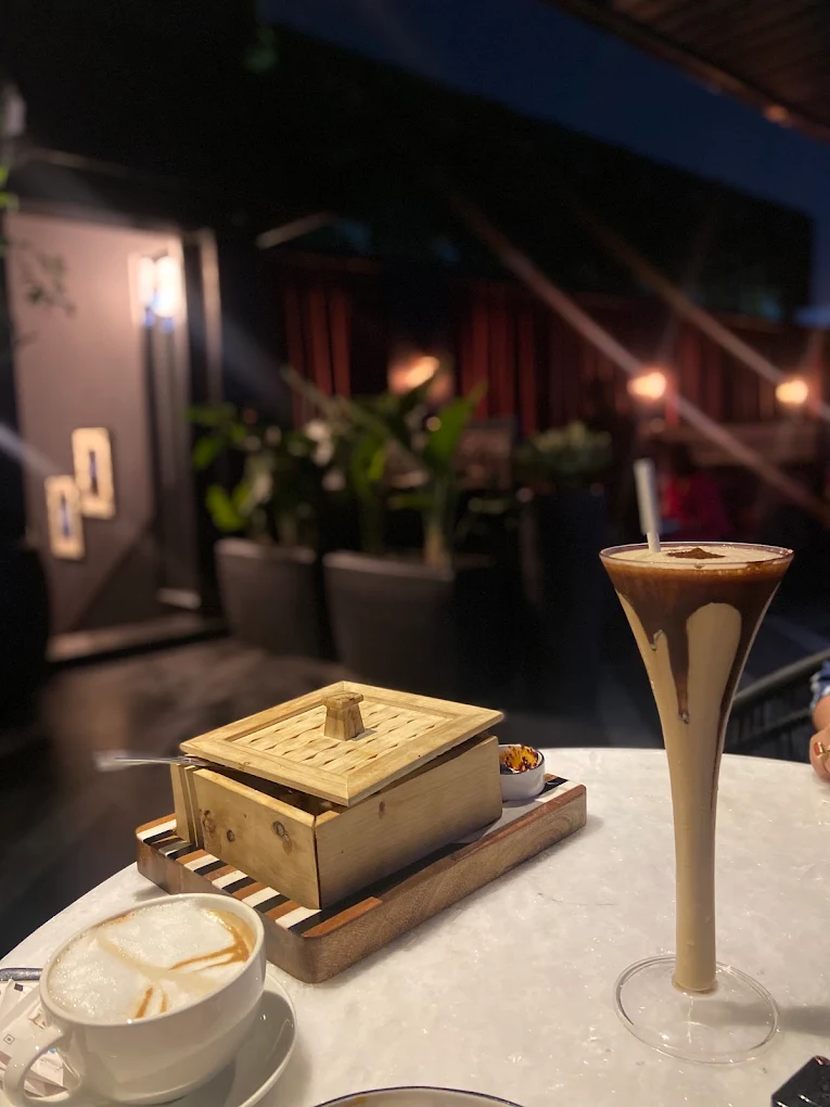 Cold coffee and cappuccino on a rooftop table at dusk, plants behind.